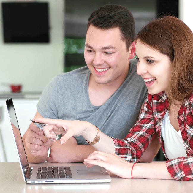 Couple sitting at a table in front of a laptop. She's pointing to the screen and smiling because they're buying a Home - Oliver & Co. Conveyancing (1)