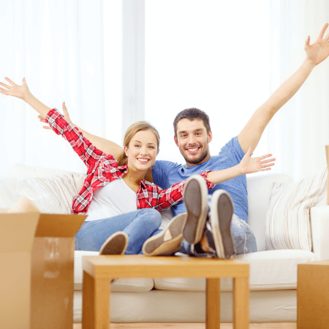 Couple on a couch with moving boxes to the side but their hands are in the air because they are Buying a Home - Oliver & Co. Conveyancing (2)